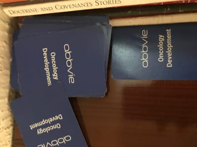 The image displays multiple blue rectangular cards with rounded corners, each printed with the white text "abbvie" in a larger, lowercase font, and "Oncology Development" stacked below in a smaller, sans-serif font. A substantial stack of these cards is located in the upper-left portion of the frame. Several individual cards are scattered on a dark brown, reflective surface, which occupies the lower and right sections of the image. One card is prominently positioned in the lower-left, rotated approximately 90 degrees counter-clockwise relative to the text on the main stack. Another single card lies flat in the upper-right, oriented similarly to the stack. A textured, light-colored material, possibly a blanket or rug, is visible along the far-left edge. Across the top, a dark, thin horizontal object is present, above which a book spine with the partial text "DOCTRINE AND COVENANTS S" is discernible. No individuals are depicted. The scene is located in Calabar, Nigeria.