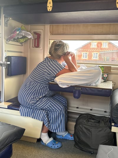 A medium shot captures a woman with blonde hair, wearing a blue and white striped dress and blue Crocs, looking out a window on a train in Shemordan, Russia. She is seated on a blue bench with her hands clasped near her chin. A white pillow and a small box are on a tray table in front of her. To her right, a black duffel bag rests on the floor. The train car interior is visible with overhead compartments containing items and a red object in a wall compartment. Outside the window, a brick building with several windows can be seen.