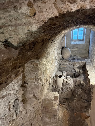 This image depicts a subterranean archaeological excavation site within the city of Cádiz, Spain, specifically consistent with the Yacimiento Arqueológico Gadir. The scene is characterized by ancient, roughly hewn stone walls and a prominent vaulted ceiling, indicating significant historical construction.

In the foreground, an archaeological pit reveals layers of earth and rubble. Partially embedded within this excavation is a large, light-colored ceramic amphora, oriented vertically with its rim visible. Further into the background, beneath another section of the stone archway, a second, similarly large, light-colored ceramic amphora stands upright on a raised platform or ledge of excavated material.

Beyond this second amphora, a distinct archway frames a distant, rectangular, multi-paned window. Light blue, translucent curtains obscure the view through this window, which illuminates a section of a plain, light-colored wall beyond. The ground level throughout the depicted area is uneven, showcasing exposed soil, stones, and remnants of older structures. No people are visible in the scene. The overall environment appears to be an active or preserved archaeological display, lit to highlight its historical features.