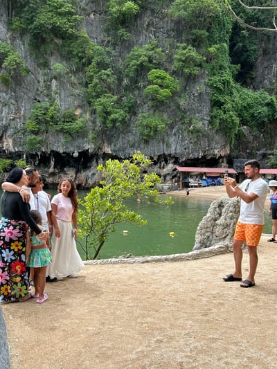 A group of tourists are gathered on a sandy path in front of a body of green water. The water is bordered by a rocky cliff face covered in lush green vegetation. In the background, several small buildings with red roofs line the water's edge.
On the left of the image, a woman wearing a black long-sleeved top and a colorful floral skirt stands with her arm around a man. They are joined by two children and another woman in a pink shirt and white skirt.
On the right, a man in an orange patterned shorts and a white t-shirt is taking a photo with his phone. Another person, partially visible, stands behind him. The overall scene suggests a leisurely day at a scenic location, likely a tourist attraction in the Krabi region of Thailand, known for its dramatic karst formations and clear waters.