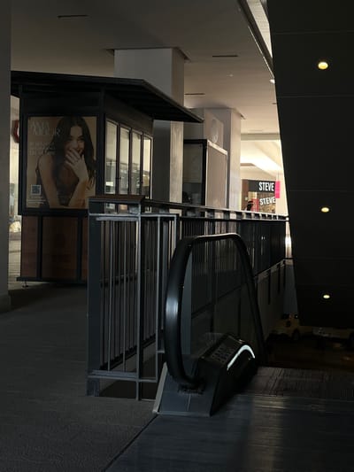 An interior shot of a modern building, featuring escalators with black railings. The lower level includes large advertisements with an image of a woman and text in Spanish. Higher up, there's a "STEVE" storefront, suggesting a retail space. The architecture is contemporary, with geometric shapes and neutral color tones. The lighting is a mix of natural light and artificial ceiling lights, creating a contrast between brighter and darker areas.