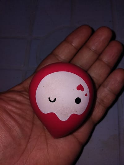 A close-up shot shows a person's hand holding a small, red, egg-shaped toy. The toy has a white face with a winking eye, a black dot for a nose, and two small red hearts above the nose. The background is a dark, textured surface, possibly a wall or floor. The image does not provide any information about the location in Haveli Jhangranwali, Pakistan.