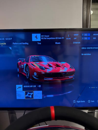 The image depicts a video game screen. It showcases a 2011 Ferrari #62 Risi Competizione 458 Italia GTC, with options to modify it. The current performance index (PI) is 832 R, with a limit of 900 R. Car level is 1. The 'BUILD RESTRICTION' shows "No restrictions." Options available include tires, wheels, drivetrain, and adjustments to "Platform and Handling" and "Fuel and Air". A highlighted option reads "INCREASE AIR FLOW". The game interface includes "Toggle Details" and "Back" buttons. The screen is being viewed through a steering wheel.