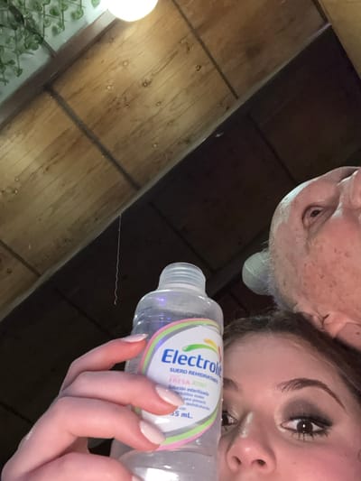 A woman holds a bottle of Electrolit in her right hand. Part of an older man's face is also in the frame. The bottle is clear with a white and blue label. The woman has manicured nails with a light pink polish. The background consists of a paneled wooden ceiling and a plant in the corner. The perspective is from below, looking upward.