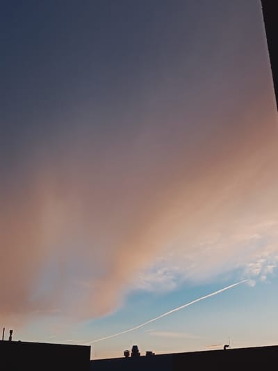 This image captures a dusky sky over a building in Peetri alevik, Estonia. The sky transitions from a deep indigo at the top to lighter shades of purple and orange as it nears the horizon, indicating either sunrise or sunset. A large, wispy cloud formation dominates the upper and middle portions of the frame, tinged with warm, peachy hues. Below this, a clear blue sky is visible, streaked with a bright white contrail left by an airplane. The lower section of the image is silhouetted by the rooftops of buildings. Visible on the rooftops are architectural elements such as chimneys and antenna-like structures. The overall scene evokes a sense of calm and the quiet transition of day. There is no visible text in the image.