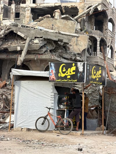 This image captures a street scene in the city of Jabalia, Palestinian Territory, depicting a makeshift retail operation in front of a severely damaged multi-story building.
The background is dominated by the remnants of a concrete structure, likely a residential or commercial building, exhibiting extensive destruction. Large sections of concrete slabs, twisted rebar, and general rubble are haphazardly piled, indicating a partial collapse. Visible architectural elements on the right side include arched windows and decorative stonework on a less damaged section of the facade, with a solar panel array mounted on its rooftop. Electrical wiring, including red and blue conduits, hangs loosely from the damaged framework. The ground leading up to the building is covered in debris, dust, and scattered refuse.
In the mid-ground, a temporary stall, possibly a kiosk, stands directly in front of the damaged building. This stall is constructed from white tarpaulin or fabric stretched over a simple frame, providing a small, enclosed space. Above the stall, two black banners with yellow Arabic script and imagery are displayed. The text "غيث 2" (Ghaith 2) is prominent. One banner features a hand holding a lit cigarette emitting smoke, while the other shows a dark, smoky image. Below the primary text, the phrase "لجميع أنواع السجائر والمعسل" (For all types of cigarettes and tobacco/ma'assel) is visible.
Three males are present around the stall. One male, wearing a dark vest over a dark long-sleeved shirt and dark shorts/trousers, stands slightly inside the stall's opening, leaning forward with his right arm extended towards items displayed on a counter within the stall. He appears to be barefoot or wearing sandals. Behind and to his right, a second male in a brown shirt is partially obscured. A third male is visible further to the right, mostly obscured by the second individual. The stall's counter displays various small bottles, packages, and other merchandise.
An orange mountain bicycle with black tires and front suspension is parked upright directly in front of the stall, facing left. The ground in the immediate foreground is uneven, consisting of packed dirt, small stones, and scattered debris. The sky above is overcast.