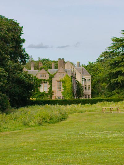 The image shows a large, old house, possibly a historic manor, situated in a rural or park-like setting. The house is made of stone and has a distinctive architectural style with multiple gables and chimneys. Ivy is growing up the sides of the building, adding to its aged appearance. The house is surrounded by lush green trees and a manicured lawn. In the foreground, a picnic table and benches are visible on the grass. The sky is overcast, suggesting a cloudy day. This property is located on the outskirts of Rugby, United Kingdom, and appears to be a well-maintained estate, likely a tourist attraction or a private residence with extensive grounds. No people or animals are present in the image, and there are no ongoing actions or interactions depicted.