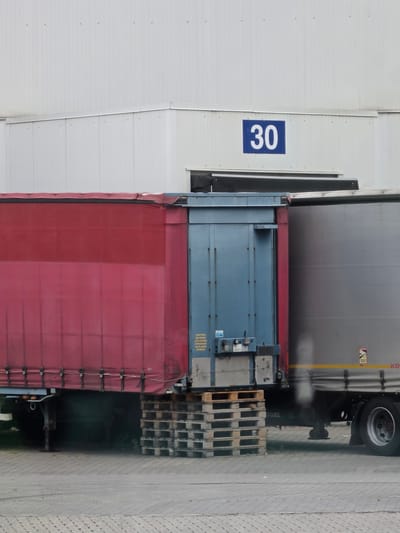 The image shows the exterior of a building with a large sign displaying the number "30". In front of the building, two semi-trailer trucks are parked. The truck on the left has a red tarpaulin cover, and the truck on the right has a grey metal trailer. The scene appears to be a loading dock or industrial area, possibly at the Frankfurt Airport in Zeppelinheim, Germany, given the context of the location. There are no people visible in the image.