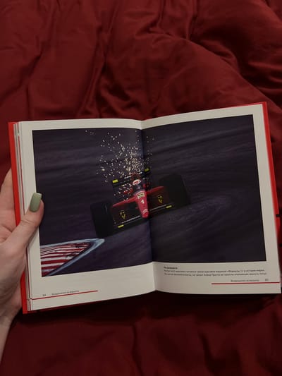 A hand with a light green nail polish is holding open a book with a red cover. The book displays a full-page image of a red Formula 1 race car with sparks flying from its rear. The car is on a race track with a curved section in the foreground marked by red and white stripes. The background is dark, suggesting nighttime or a dimly lit environment. Text is visible at the bottom of the open pages. The left page has the number "84" and the Russian text "Возвращение на вершину" (Return to the top). The right page has a paragraph of Russian text, followed by the phrase "Возвращение на вершину" and the number "85". The image does not contain any information about the city of Vysokaya Gora, Russia.