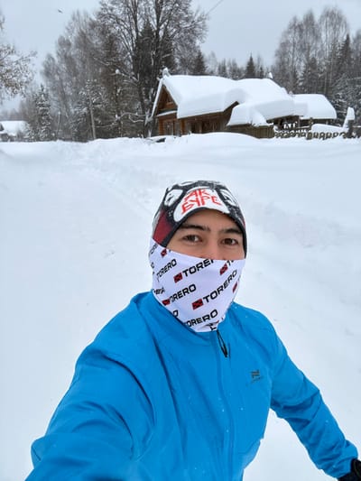 A person in a blue jacket and a balaclava stands in snowy terrain, with a building covered in snow visible in the background. The person's face is partially covered, leaving only their eyes visible. Snow covers the ground and surrounding landscape. The overall scene suggests a cold, wintery environment.