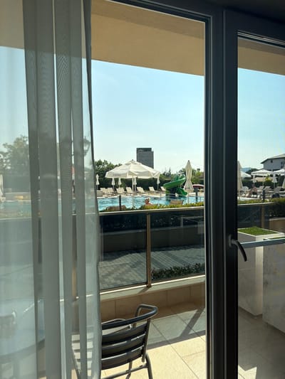 This image captures a view from a room overlooking a resort pool area in Hisarya, Bulgaria. The foreground is dominated by sheer white curtains on the left and a glass door frame on the right. Beyond the glass, a bright blue swimming pool with lounge chairs and umbrellas is visible. To the left of the pool, a green water slide is prominent. Several white umbrellas are scattered around the pool area, with one bearing the "EMPIRE" logo. In the background, a multi-story building and trees can be seen under a clear blue sky. A few individuals are present by the pool, some relaxing on lounge chairs, and one person is in the water. The balcony area visible through the glass features a patterned tile floor and a metal chair.
