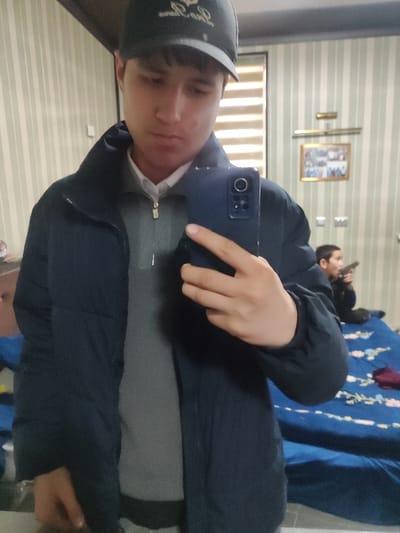 The image displays a mirror selfie captured indoors in a room within the city of Rishtan, Uzbekistan. The primary subject, a young male, is visible from the chest up, wearing a dark baseball cap with a light-colored logo, a dark blue puffer jacket, a light gray zippered sweater, and a white collared shirt. He holds a dark blue smartphone with his right hand, covering the lower portion of his face and central torso. His facial expression is neutral. The reflected background features a wall with vertical light-colored stripes. A framed picture, illuminated by a small gold-toned wall light fixture, hangs on the wall. To the right of the primary subject's reflection, a younger male is seated on a bed. This individual is dressed in dark clothing and holds a dark object resembling a pistol or toy gun, pointing it away from the mirror. The bed is covered with a dark blue blanket or sheet featuring a light-colored floral or patterned design, and a dark red fabric item is also present on the bed.