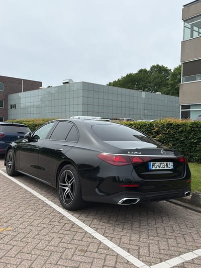 A black Mercedes-Benz E220d sedan is parked in a designated parking space on a brick paver surface. The car's license plate is HG-403-NJ. Behind the car is a dense hedge. Further in the background, there is a modern building with a glass facade. To the left of the Mercedes, a dark blue SUV is parked. The sky is overcast. The image was taken in the city of Son en Breugel, Netherlands.