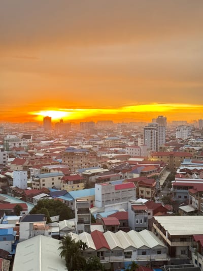 This media file captures an aerial view of a dense urban landscape in Khan Mean Chey, Cambodia, bathed in the warm, orange glow of a sunset. The sky is dominated by an expansive, brightly lit horizon, with the sun appearing as a luminous orb partially obscured by wispy clouds. The atmospheric conditions suggest a clear, possibly humid evening.

The scene is characterized by a sprawling collection of buildings, predominantly residential, with a mix of architectural styles and roof colors, including red, white, blue, and grey. Many structures are multi-story, closely packed together, creating a textured urban fabric. Some buildings feature balconies, while others appear to be more utilitarian in design. Glimpses of greenery, including palm trees, suggest pockets of vegetation within the built environment.

The time of day is clearly sunset, indicated by the intense coloration of the sky and the long shadows that would be cast if the perspective were horizontal. The overall mood conveyed is one of a vibrant, densely populated urban area at the close of day, perhaps a moment of transition from work to evening. No people are distinctly visible, and the focus is on the cityscape and the dramatic sky. No visible text is present in the image.