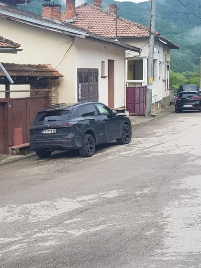 In Lomets, Bulgaria, a dark-colored Volkswagen SUV is parked on a paved street in front of a light-colored building with a red-tiled roof. The vehicle, which has a license plate reading "PA 5804 MB", is positioned at an angle, facing away from the viewer. A second, black SUV is visible further down the street, also parked. The environment appears to be a residential area, with houses and a mountainous backdrop. The road surface shows signs of wear and tear, with patches of lighter-colored material. A wooden fence and a bench are present to the left of the Volkswagen. The sky is overcast.