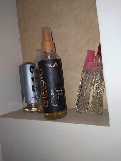 The image depicts five personal fragrance bottles arranged on a white shelf within an interior space. The rear wall is light beige with a subtle texture, while a white vertical partition borders the right side of the shelf. From left to right: a cylindrical bottle with a silver-colored upper section and a clear glass base containing a light yellow liquid, partially obscuring a smaller, clear glass bottle with a transparent spray top; a central opaque black spray bottle with a gold-colored sprayer, labeled "MAGIC Perfumes Body Splash" and "Body," containing a clear yellowish liquid; and two slender, clear glass bottles with faceted or ridged textures and pink spray tops, containing clear liquid. The leftmost bottle displays partial text "SWE". The precise location within the city of Harasta, Syria, is not discernible from the visual content of the image.