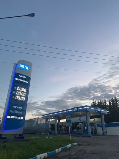 The image shows a gas station in the city of Vereya, Russia. A tall blue and white sign displaying fuel prices and the brand "ОПТИ" (Opti) stands prominently on the left. The sign shows prices for 95, 92, and D (diesel) fuel. Below the fuel prices, the words "Магазин" (Shop) and "AdBlue" are visible. The gas station canopy, also branded with "ОПТИ", is lit with blue lights. Several fuel pumps are visible under the canopy. A sign indicating "ОБЯЗАТЕЛЬНАЯ ВЫСАДКА ПАССАЖИРОВ" (Mandatory Passenger Drop-off) is also present. The sky is overcast with clouds, and a faint crescent moon is visible. There are power lines stretching across the sky. Trees line the background of the gas station. The ground is paved asphalt with some grassy areas.