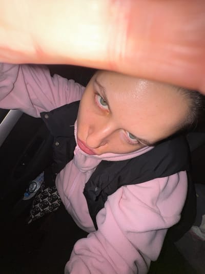 A high-angle selfie depicts a person with light skin, light eyes, and dark hair. They are wearing a pink hooded sweatshirt under a black jacket. Part of a hand or arm is visible in the foreground, partially obscuring the left side of the frame. Below the person, parts of patterned black and white pants and a plastic bottle can be seen, along with a dark surface. The exact location in Uvarovo, Russia cannot be determined from the image.