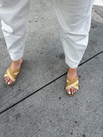 This is a top-down, eye-level shot of a person's legs and feet from the knees down, standing on a textured concrete surface. The person is wearing white pants and woven slide sandals. The sandals are a criss-cross design in neutral beige and tan colors, with a cork sole. The toes are painted a light mint green color. A thin, black line runs diagonally across the concrete in the foreground, separating two sections of the sidewalk. The concrete itself has a rough, speckled texture with various shades of gray and small stones. The lighting suggests it is daytime, likely sunny given the bright white of the pants and the crisp shadows. The scene appears to be outdoors, on a sidewalk or paved walkway. There is no visible text in the image.
