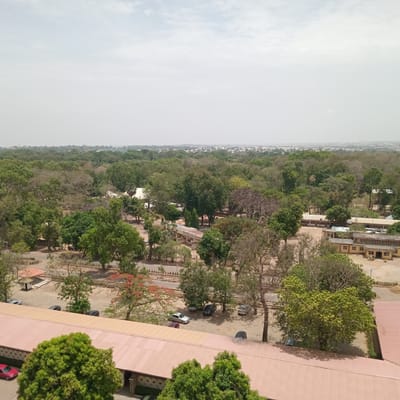The image depicts an aerial view of an urban landscape in Jama A, Nigeria. A dense canopy of trees dominates the midground, interspersed with buildings, primarily featuring light-colored walls and red or brown roofs. In the foreground, a series of connected structures with red roofs are visible, partially obscured by greenery. A parking area adjacent to these buildings contains several parked cars. In the background, the city extends towards the horizon, characterized by a mix of vegetation and structures under a slightly overcast sky.