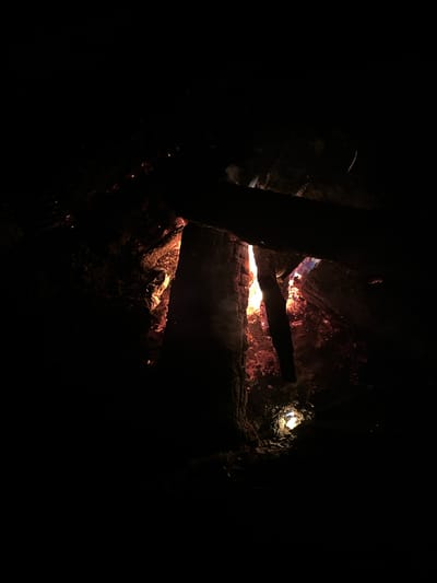 The image depicts a dark scene dominated by a fire. Burning wood and embers are visible within what appears to be a fire pit or contained area. A few logs are positioned over the flames, creating shadows and obscuring the intensity of the fire. The surrounding environment is extremely dark, making it difficult to discern details beyond the immediate vicinity of the fire. A single light source, possibly a lamp, illuminates the rock walls, far right. The location is Юзуфоўскі сельскі Савет, Belarus.