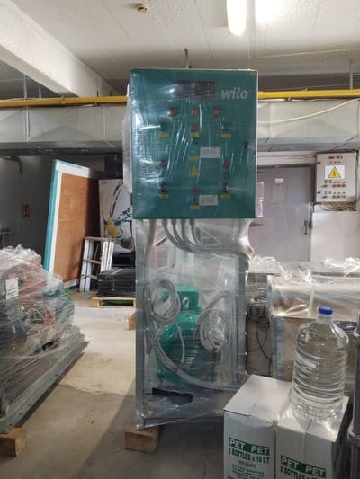 The image shows an indoor scene with various objects and equipment. In the foreground, there's a piece of machinery with the brand "Wilo" visible. It's wrapped in clear plastic, possibly for protection or packaging. Next to it, there are boxes of PET water bottles, stacked with a clear water bottle on top. Text on the boxes indicates "PET PET 2 BOTTLES x 10 LT" with Greek text below and "ZAROS KRHTHS", which seems to indicate it is from Crete.

Behind these objects, there's a brown table-like surface with a white and teal border. Further back, there appears to be more machinery covered in plastic.

The background includes a gray wall with an electrical panel containing a yellow triangular warning sign. There's also a closed gray metal door and various conduits and pipes running along the wall.

Based on the text "ZAROS KRHTHS," which translates to "Zaros Crete," the image appears to be taken in Greece, possibly in or near the Zaros region. The location in the city of Pyli, Greece, is not explicitly mentioned but cannot be ruled out. The objects and equipment suggest an industrial or commercial setting.