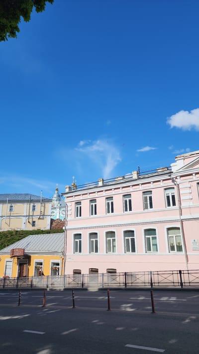 The image depicts a street scene in Smolensk, Russia. A pink building with multiple windows occupies the right side of the frame, alongside a road with traffic barriers. Adjacent to the pink building, there is a beige building with a dome topped with crosses. A yellow building with a sign is also visible. The left side of the image is dominated by a clear blue sky with scattered clouds.