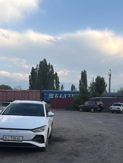 The image shows an outdoor scene with several vehicles and shipping containers. In the foreground, a white car with the license plate "01 736 BC" is parked. Behind it, a dark-colored SUV with "TOYOTA" visible on the rear is parked, followed by a blue shipping container with Cyrillic text that appears to read "БАЛТІ" and some numbers. Further back, a white car is parked. The scene is set in a lot with a gravelly surface, and trees and mountains are visible in the background. The sky is partly cloudy with bright sunlight, suggesting it is daytime, likely late afternoon due to the warm light. There are no people visible in the image. The overall impression is one of an industrial or transportation hub.