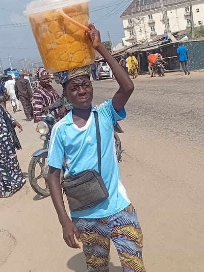 A man in a light blue shirt and patterned pants balances a large container of what appears to be food on his head. He is also wearing a dark crossbody bag. He stands next to a motorcycle on an asphalt surface, possibly a road. In the background, there are buildings, other people, and various objects that suggest a bustling outdoor setting. The image is slightly rotated, giving a tilted perspective.