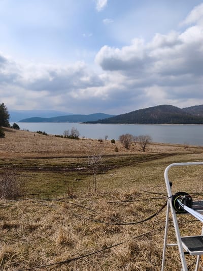 The image depicts a wide outdoor landscape located in the city of Sv. Konstantin, Bulgaria, under a partly cloudy sky. The foreground features a gently sloping terrain covered predominantly with dry, yellowish-brown grass and some patches of darker, possibly moist, soil. In the lower right quadrant, a white, metal step ladder is visible, with a black cable spool or enclosure mounted on one of its steps. Black cables extend from this equipment, running horizontally and diagonally across the dry grass in the foreground.
The midground transitions to a large, calm body of water, likely a reservoir or lake, stretching horizontally across the scene. The immediate shoreline is sparsely populated with small, leafless deciduous trees and bushes. Beyond the water, a series of dark green, forested hills and mountains rise, forming the distant background. The sky above is a mix of light blue patches and prominent grey and white cumulus clouds. No people are visible. The overall impression is one of late winter or early spring, characterized by dormant vegetation.