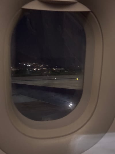 The image shows a view from an airplane window at night. Outside, city lights are visible. The wing of the plane is also visible through the window. It is impossible to determine if the location is Churriana, Spain from the available image.