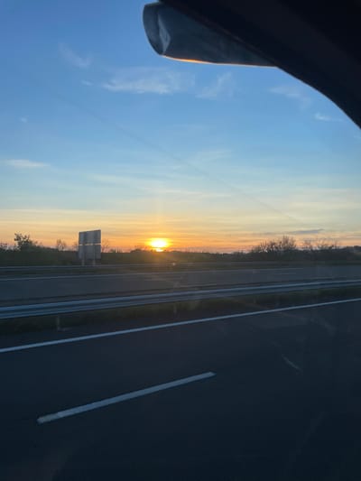 The image presents a highway scene at sunrise or sunset, observed from within a vehicle, in the vicinity of Portel-des-Corbières, France. The sky, occupying the upper two-thirds of the frame, exhibits a gradient from clear blue at the zenith to vibrant orange and yellow hues at the horizon. The sun is a prominent, bright orange disc positioned low on the horizon, emitting a significant glare and lens flare across the lower sky. Wispy clouds are scattered, and a faint contrail stretches diagonally across the upper portion of the sky.
A dark, curved structure, identified as an interior component of the viewpoint vehicle (e.g., sun visor or mirror housing), partially obscures the upper right and central top areas of the frame. The foreground shows a multi-lane asphalt highway with clearly delineated white lane markings: a solid line along the left edge of the visible road and a dashed line positioned closer to the viewer. A metal guardrail, comprising two horizontal silver rails, runs parallel to the road, separating the immediate lanes from adjacent highway segments. Beyond this guardrail, additional dark asphalt lanes are visible.
The environment bordering the highway consists of various green trees and shrubs. On the left side of the highway, past the opposing lanes, a large, rectangular, light-colored highway sign structure stands. No individuals or other vehicles are discernible within the frame. The scene is illuminated by the direct, low-angle sunlight.