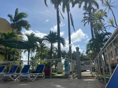 The image depicts a scene at a resort or hotel in Le Gosier, France. Several palm trees are visible against a blue sky with scattered clouds. In the foreground, there are lounge chairs with blue cushions arranged along a tiled patio or walkway. A metal railing and signage are also present, with text indicating “Plage” (Beach), “Snack”, and “Restaurant”. The scene appears to be near a pool or beach area, with tropical vegetation framing the view.
