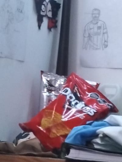 A casual image showcases a cluttered space. A red bag of Doritos rests atop a pile of clothing and possibly a book. Behind it is another silver Doritos bag. The background includes drawings or posters on a white wall. One drawing appears to depict a soccer player in uniform. A dark pole or rod is positioned vertically in the frame, behind the bags of chips. The scene gives off a relaxed and somewhat messy impression.