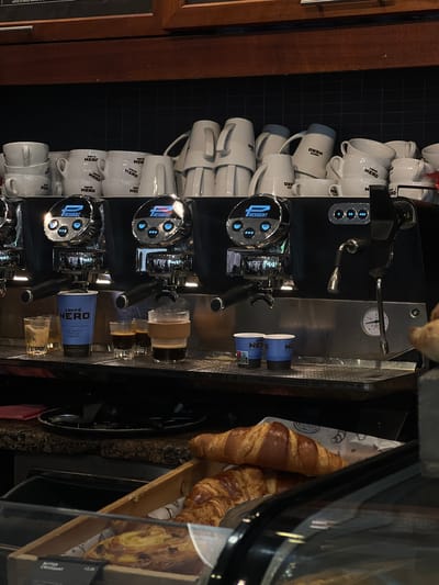 The image shows a coffee preparation area, likely inside a Caffè Nero branch, possibly in Belfast, United Kingdom given the branding on the coffee cups and espresso machines. The counter features multiple Resident espresso machines with coffee cups stacked on the side. There are also croissants and other pastries in a display case adjacent to the coffee making area. The scene is brightly lit, with a dark background suggesting it is taken in a commercial setting. No people are visible in the frame.
