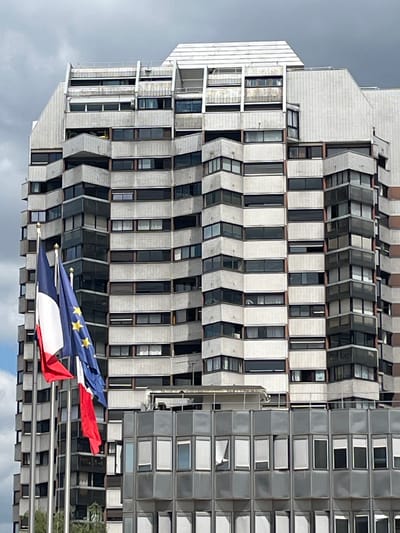 The image shows a modern, multi-story building with a distinctive stepped facade and numerous windows, some with balconies. In the foreground, three flags are visible: the French flag, the European Union flag, and another flag, possibly red. The flags are on flagpoles, with the French flag and the EU flag prominently displayed. The building appears to be in an urban setting, with a cloudy sky overhead. This building is located in Créteil, France.