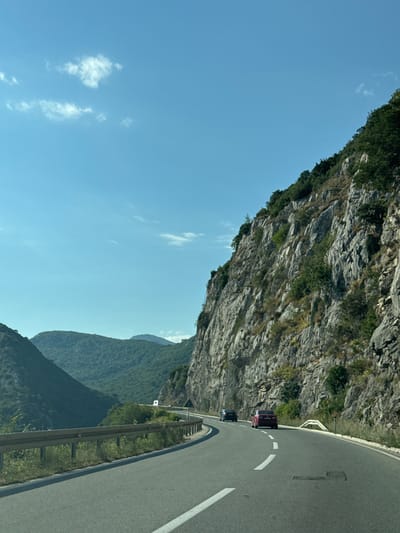 The image depicts a scenic road winding through a mountainous landscape. The road surface is asphalt, marked with white lines. A metal guardrail runs along the left side of the road, bordering a steep, rocky cliff face covered in sparse vegetation. In the distance, rolling green hills and mountains are visible under a bright blue sky with scattered white clouds. Several cars are present on the road: a dark-colored car and a red car are in the foreground, with another vehicle further ahead. The scene appears to be a travel route in a natural, undeveloped area. The location is identified as Košljun, Montenegro.