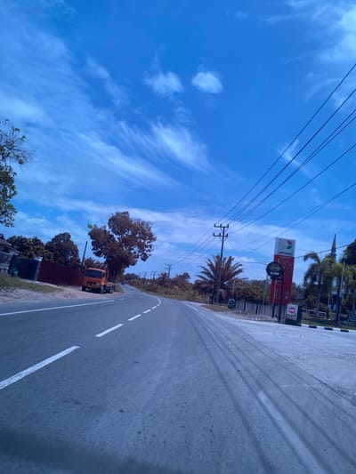 This image depicts a street scene in Aluepiet, Indonesia, under a bright blue sky with wispy white clouds. The road curves to the right, with white lane markings visible. On the left side of the road, a red fence separates the street from residential buildings and lush green trees. An orange truck is parked on the shoulder. On the right side of the road, a Pertamina gas station sign with a red and white design is prominent, displaying prices and the text "24 JAM". Power lines stretch across the upper portion of the image, supported by utility poles. Palm trees and other vegetation line the side of the road. The overall impression is that of a somewhat rural or suburban setting on a clear day.