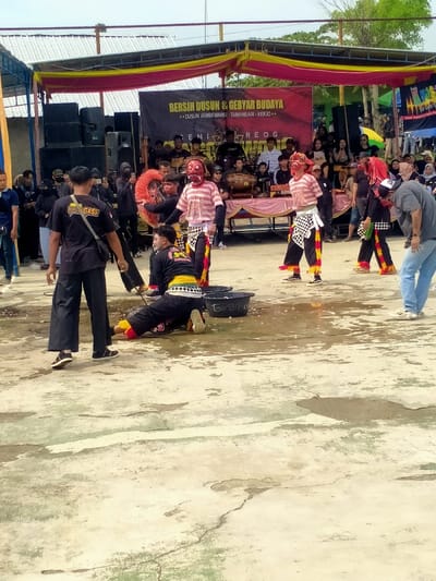 An outdoor cultural celebration is taking place in Dusun Jambewang, Tamansari, Kerjo, Indonesia. The scene is set on a light-colored, cracked concrete or paved ground with visible moisture and small puddles. In the foreground, approximately six individuals are engaged in a performance. Three central figures wear red, demon-like masks, striped red and white shirts, and black pants with vibrant red, yellow, and white/black checkered patterns; one holds a red feathered fan-like prop. To the left of these masked performers, an individual in a black shirt with yellow trim and black pants is prone on the ground, facedown on hands and knees, positioned next to a black bucket labeled "Gudang". Another person stands nearby, wearing a black t-shirt with "GASS GASS" on the back and loose black pants, observing the prone performer. On the far right, a man in a grey t-shirt and blue jeans leans forward, watching the performance.
In the mid-ground, a raised stage, draped with pink fabric, hosts musicians playing traditional instruments, including drums and gongs. Large black speaker stacks flank both sides of the stage. A prominent black banner stretches across the stage, displaying the Indonesian text "BERSIH DUSUN & GEBYAR BUDAYA" (Village Cleaning & Cultural Celebration), along with "DUSUN JAMBEWANG - TAMANSARI - KERJO" and "GENI REOG SINGO MUDHO". The entire stage area is partially covered by a temporary metal-frame canopy draped with yellow and red fabric. A crowd of people, varied in age and attire, is visible behind the stage, observing the event. The scene is brightly illuminated by natural daylight, with a pale sky overhead.