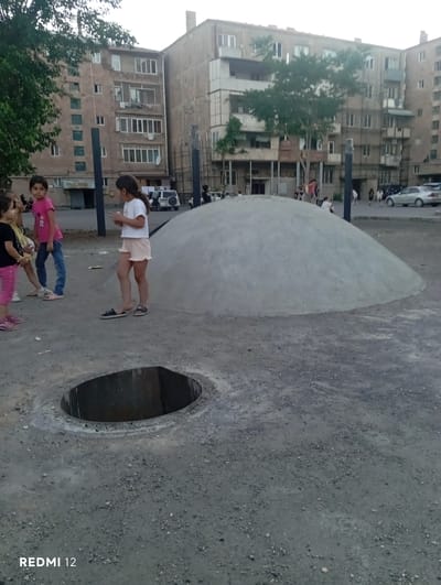 In an outdoor urban environment, likely a playground or open space in Vedi, Armenia, a group of young girls are observed. The scene features a large, smooth, dome-shaped concrete structure in the mid-ground. To the left, three girls are visible; one in a pink t-shirt and blue jeans, another in a white t-shirt and light-colored shorts, and a third in a black t-shirt and pink pants. A fourth girl, wearing a white t-shirt and light-colored shorts, stands near the dome. In the background, apartment buildings dominate the skyline. A dark, circular opening, possibly a manhole or drain, is in the foreground. Cars and other people are visible in the distance, suggesting a public area. The lighting indicates it is likely late afternoon or early evening.