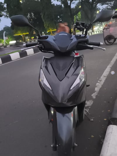 The image captures a dark gray Honda scooter positioned on the side of an asphalt road. The scooter is viewed from the front, highlighting its sleek, angular design, prominent headlights/turn signals, and front fender. The word "HONDA" is distinctly visible in white lettering on the scooter's front fairing. A phone holder or mounting device is attached to the handlebars, and a plastic bottle with a red cap is partially visible in a storage compartment beneath the handlebars. The scooter's mirrors are extended.
The immediate surroundings include a black and white striped curb or road divider running parallel to the road on the left, and a white, partially faded line on the asphalt to the right. The road surface appears dark, characteristic of asphalt. In the background, numerous trees with dense foliage line the road, and several streetlights are illuminated, casting a soft glow that suggests the time of day is dusk or early evening.
Further in the background on the right, a light-colored, wheeled vendor cart or food trolley is visible. Among the trees, two abstract, light-toned figures, possibly statues or blurry individuals, can be discerned. The overall scene depicts a quiet street setting, likely in an urban or suburban area, consistent with the Cibinong, Indonesia context. The scooter is stationary, and no specific activity or event is taking place beyond its presence on the roadside.