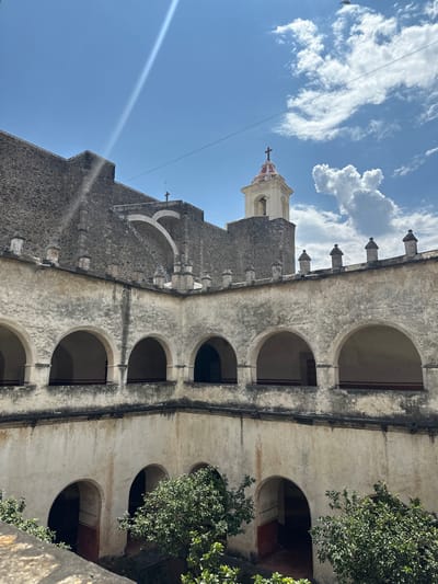 The image shows the former monastery of the Nativity of the Virgin Mary in Tepoztlán, Mexico, viewed from an external vantage point at a low angle. The architecture features stone construction with a cream and gray facade, arches, and a visible church tower topped with a cross against a blue sky with scattered clouds. Green foliage is seen at the right side of the image, framing the building.