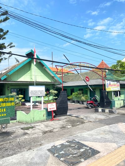The image shows the entrance to a military building in Kecamatan Tulangan, Indonesia, located at latitude -7.481 and longitude 112.645.  A red and white Indonesian flag hangs from a pole on a green building to the left of the entrance. The entrance is marked by a red and gold arch with the words "KORAMIL 0816/05 TULANGAN" inscribed on it.  The arch is flanked by two black pillars, one of which is topped with a red stop sign. A red and white sign on the right side of the entrance reads "POMDAM V/BRW" with a black and white  sign below it that is not legible in the photo.  There is a small, red, wheeled scooter parked to the right of the entrance.  The entrance is paved with gray concrete, which extends outwards in a slight downward slope. There are several green plants and trees surrounding the building. The sky is blue with white clouds.  The building has a green painted facade and a brown roof.  A sign on the left side of the building is visible with the text "PERSIT KARTINI CABANG KRAMAT RANTING 5 KORAMIL 0816/05 TULANGAN KOMANDO KEHIDUPAN NO. 31 TULANGAN".
