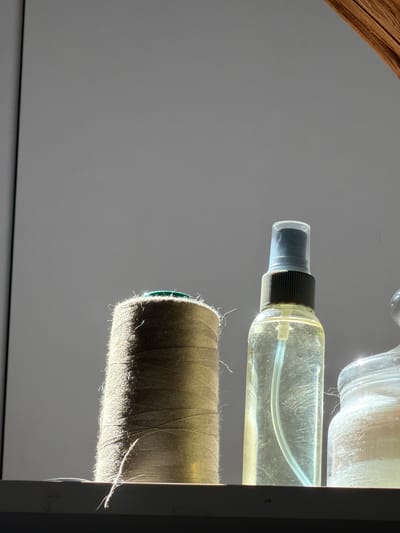 The image shows a close-up, side view of several objects on a light gray background. In the foreground, a spool of thread is visible. The thread is a light brown or khaki color, and the spool has a green edge. The thread is lit from the side, creating highlights and showing individual fibers. Above the spool of thread is a clear spray bottle filled with a light yellow liquid. The bottle has a black cap and a white straw inside. The liquid inside the bottle is slightly cloudy, with some visible particles or streaks. In the background, a portion of a clear glass jar is visible, also filled with a cloudy substance. The jar is lit from above, creating bright reflections. The overall impression is one of a collection of items, possibly related to crafting or beauty, arranged for display or use. The lighting suggests a studio setting or a deliberate arrangement to showcase the textures and transparency of the objects. There is no indication of people, specific time of day, weather, or emotions. No text is visible in the image.