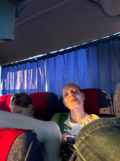 The image shows two people in what appears to be a vehicle, possibly a bus or van. The background includes window blinds and parts of the vehicle's interior. One person is awake and looking at a mobile phone, with their tongue slightly out. The second person appears to be sleeping. The location in Simakovo, Russia is not explicitly confirmed in the image, and is only specified within the request.