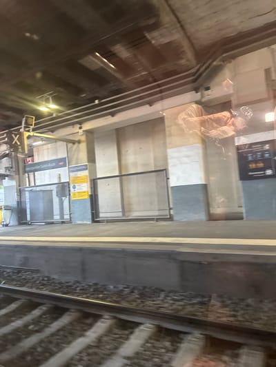 The image captures a view of a train station platform in Montpellier, France, likely during daylight hours as indicated by the illumination. The scene is viewed through a reflective surface, possibly a window or glass partition, which creates reflections of the environment.

The primary subjects include a person wearing a patterned garment, seated and possibly looking at something off-camera. The person's face is partially obscured, but they appear to be wearing a mask. The setting is an indoor or covered train station, with concrete walls and structures visible. Train tracks are also a prominent feature, running alongside the platform.

Notable details include various signs and informational displays on the walls. One sign prominently displays "Montpellier Saint-Roch," confirming the location. Other signs contain text in French, with some appearing to be yellow hazard warnings. There are also metal grates or barriers on the platform, likely for safety or access control. Artificial lighting, in the form of rectangular fixtures, is visible, suggesting it's an enclosed or dimly lit area. The overall impression is that of a functional, modern transportation hub. The reflections suggest a busy environment, though the specific activity of the visible person is unclear.