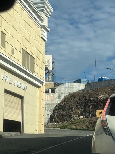 The image captures a bright, sunny day in Shkotovo, Russia. In the foreground, a light-colored building with a sign that reads "HOTEL & PARKING" is visible on the left. A large, open garage door is present on this building. To the right, a white vehicle is partially visible, showing its rear taillights. The background features a steep, rocky embankment. Construction is evident on the embankment, with scaffolding and structures partially obscuring some of the terrain. Several portable toilets are also spotted on the higher ground behind the rocky slope. The sky is a clear blue with scattered white clouds. The overall scene depicts a mix of urban development and ongoing construction in a hilly, possibly coastal, environment.