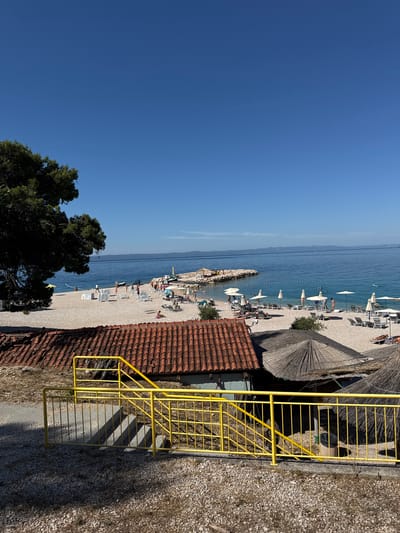 The image shows a sunny day at a pebble beach in Makarska, Croatia. In the foreground, a yellow metal railing and steps lead down towards the beach. A roof with red tiles covers a building to the left of the steps, and thatched umbrellas are visible to the right. The beach itself is dotted with lounge chairs and umbrellas, and several people are seen relaxing, sunbathing, and walking. The clear blue sea stretches out to the horizon, with a distant coastline visible. Some people are in the water, and a jet ski can be seen near a rocky breakwater. A large tree with green foliage is on the left side of the image.
