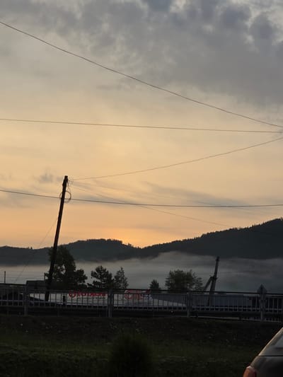 This image depicts a dimly lit outdoor scene, likely at dawn or dusk, with a soft, orange-hued sky and muted grey clouds. A power line stretches across the upper portion of the frame. In the middle ground, a dark silhouette of a forested mountain range is visible, partially obscured by a layer of fog. In front of the mountains, a metal fence runs horizontally, with several trees visible behind it. A utility pole with tangled wires stands prominently on the left side of the frame. A sign with Russian text and the number "75" is visible on a structure behind the fence. The foreground shows a dark, grassy area. The overall impression is one of a rural or semi-rural landscape with infrastructure elements.