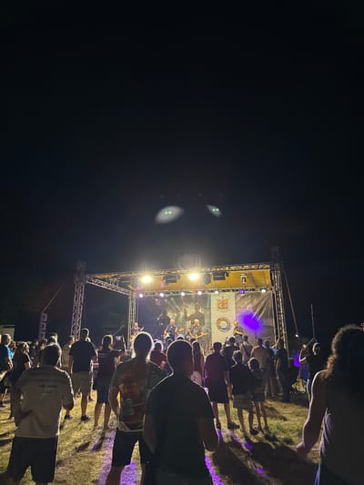 The image captures a nighttime outdoor concert scene in Tutrakan, Bulgaria. A band is performing on a raised stage equipped with lights and a large banner. The crowd, diverse in age and gender, stands on a grassy field, watching the performance. Some individuals are holding phones up, possibly recording or taking pictures. The overall atmosphere appears to be that of a lively festival or event. In the sky above the stage, two luminous green circles, suggestive of eyes, are visible, possibly an optical illusion or a deliberate visual effect.